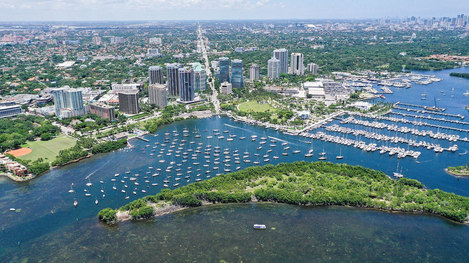 Aerial view of Coconut Grove's downtown and Dinner Key waterfront on Biscayne Bay, with sailboats and the Miami skyline beyond